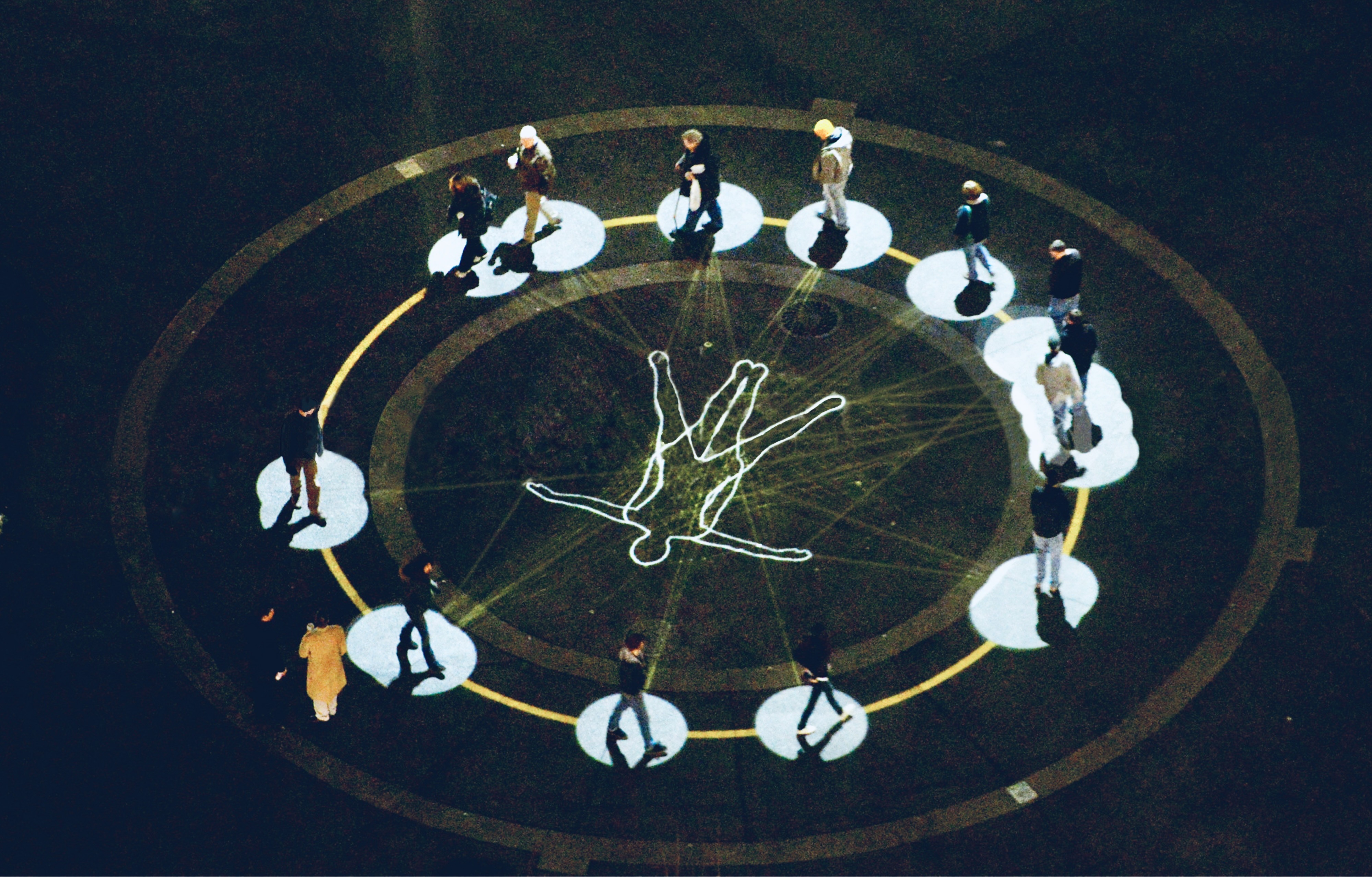 An overhead view of Congregation: a ring of people stand in individual pools of light around a public square at night, each connected by faint lines of light to a single human figure outlined at the centre.